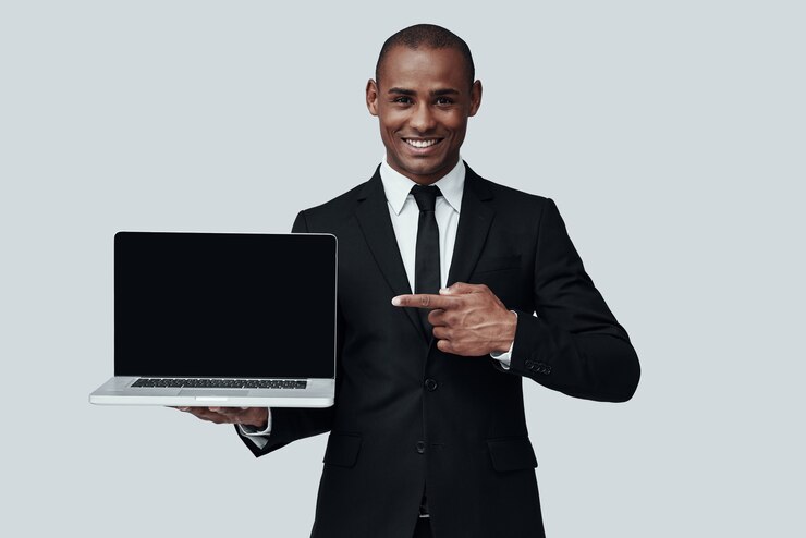Tu devrais voir ça ! Jeune homme africain en tenue de soirée pointant sur un ordinateur portable et souriant en se tenant debout sur fond gris