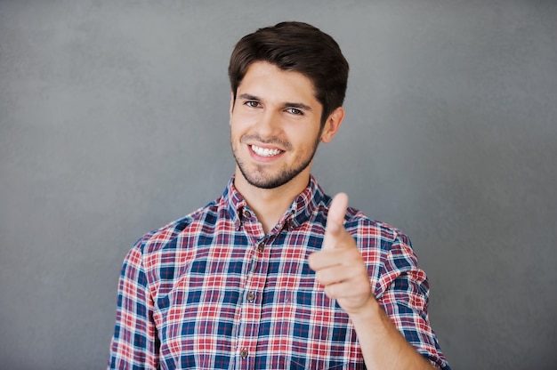 Tu peux le faire! Gai jeune homme pointant sur la caméra et souriant en se tenant debout sur fond gris