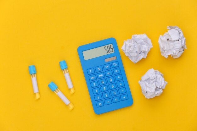 Tubes à essai, calculatrice avec boules de papier froissé sur fond jaune. Vue de dessus