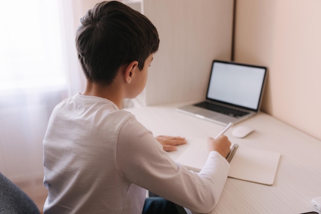 Étude d'écolier au bureau dans sa chambre Garçon utilise un ordinateur portable et écrit dans un ordinateur portable Livres et tablette sur le t