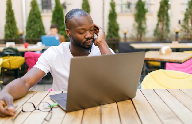 Étudiant africain fatigué portant un t-shirt blanc se reposant après avoir étudié épuisant au café en plein air