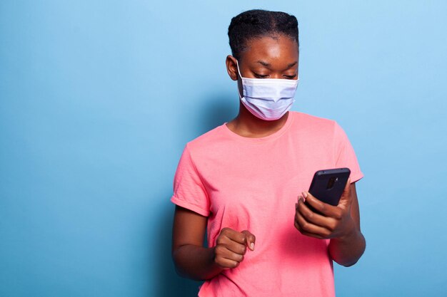 Étudiant afro-américain avec masque médical de protection contre le coronavirus tenant un smartphone naviguant sur les réseaux sociaux envoyant des SMS avec des amis en studio sur fond bleu. Texte de types de femme