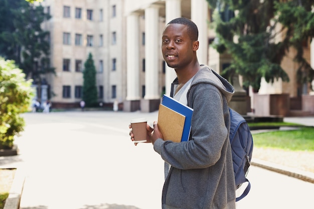 Étudiant afro-américain souriant avec des livres et du café à emporter à l'extérieur, se reposant sur le campus. Concept d'éducation, espace de copie