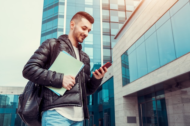 Étudiant arabe à l&#39;aide de smartphone à l&#39;extérieur. Heureux mec regarde le téléphone en face du bâtiment moderne après les cours