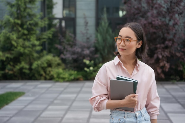 Étudiant asiatique intelligent portant des lunettes tenant un livre, un ordinateur portable debout sur le campus universitaire