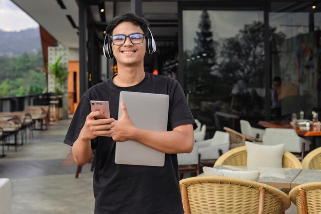 Étudiant asiatique souriant et tenant un téléphone et un ordinateur portable au café