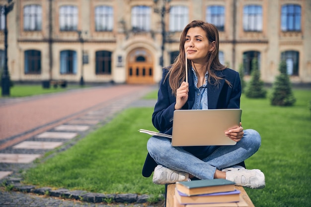 Étudiant attrayant assis sur le banc avec les jambes croisées et tenant un crayon tout en travaillant sur un ordinateur portable