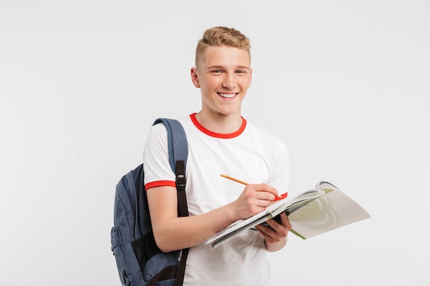 Étudiant attrayant à l'université ou au collège portant un sac à dos en souriant et en vous regardant tout en écrivant des notes au manuel isolé sur blanc