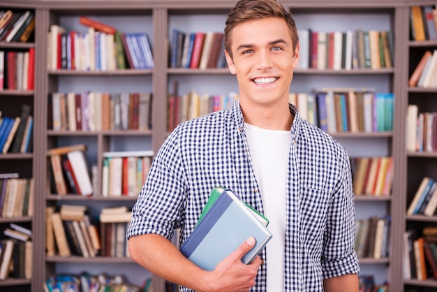 Étudiant en bibliothèque. Beau jeune homme tenant des livres et souriant en se tenant debout dans la bibliothèque