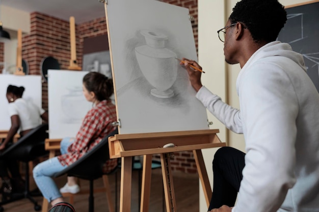 Étudiant créatif assis devant une toile blanche dessinant un modèle de vase à l'aide d'un crayon graphique travaillant à l'illustration de croquis pendant le cours d'art. Équipe diversifiée assistant à une leçon de dessin dans un studio de créativité