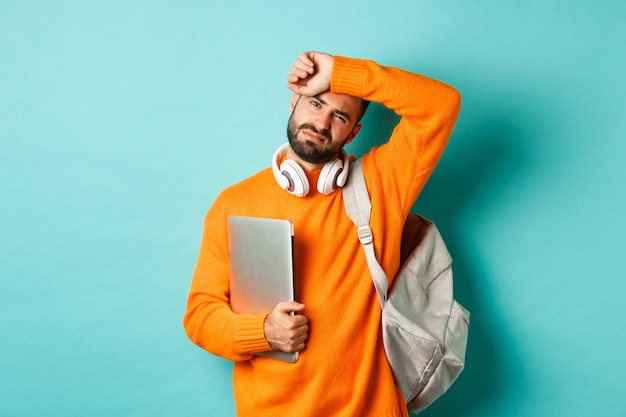 Étudiant fatigué essuyant la sueur sur le front, tenant un ordinateur portable et un sac à dos, debout dans un pull orange sur fond turquoise.