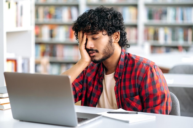 Étudiant fatigué de nationalité arabe ou indienne s'endormant à un bureau de la bibliothèque dans des vêtements élégants fatigué de se préparer à l'examen en lisant des livres se sent épuisé les yeux fermés a besoin de repos