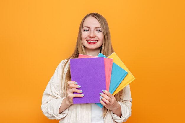 Étudiant fille blonde caucasienne en veste beige détient quatre livres dans des couvertures multicolores souriant isolé sur un mur orange. École de langue anglaise, concept d'éducation