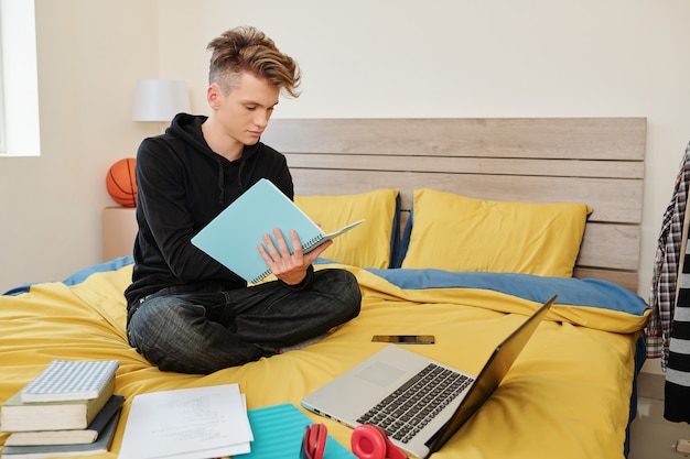 Étudiant en génie logiciel assis sur le lit avec des livres, un ordinateur portable et des manuels autour et écrit dans un cahier