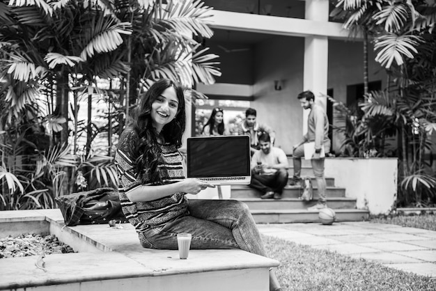 Étudiant indien asiatique concentré travaillant sur un ordinateur portable ou lisant un livre tandis que d'autres camarades de classe en arrière-plan, photo en plein air sur le campus universitaire