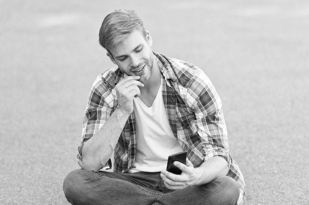 Étudiant insouciant temps libre passer du temps d'été bel homme chemise à carreaux étudiant de la mode masculine se détendre regarder un appel vidéo sur le téléphone mec macho bavarder en plein air homme de style de rue assis sur le sol