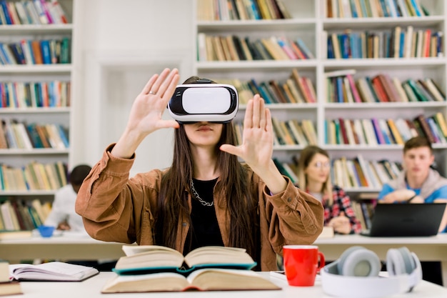 Étudiant de jeune fille brune travaillant avec simulateur VR dans la bibliothèque. Jeune femme en tenue décontractée et lunettes de réalité virtuelle assis au bureau et toucher l'air