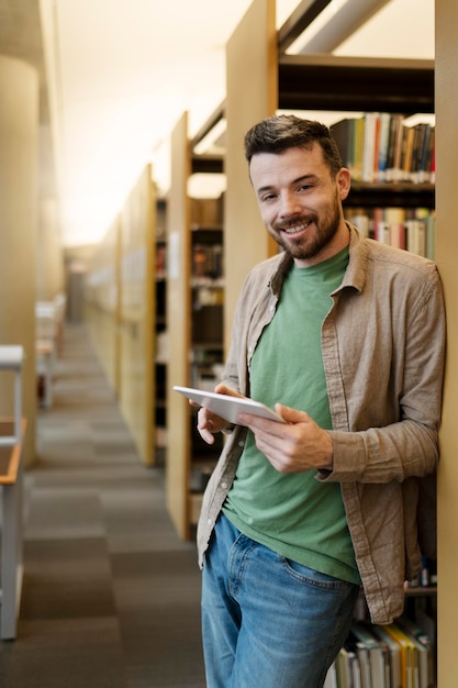 Étudiant latin souriant positif debout dans la bibliothèque universitaire à l'aide d'une tablette lisant des livres électroniques