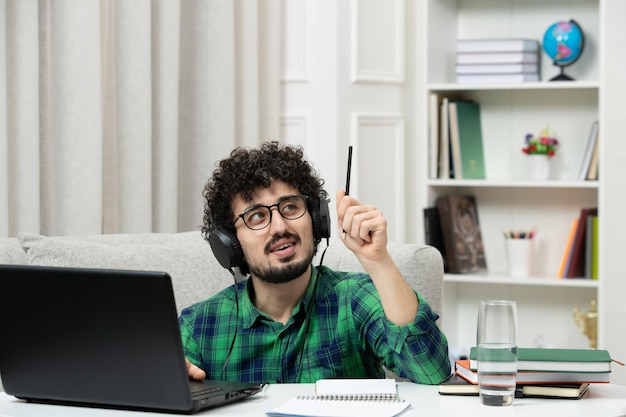 Étudiant en ligne mignon jeune homme étudiant sur ordinateur dans des verres en chemise verte levant la pensée