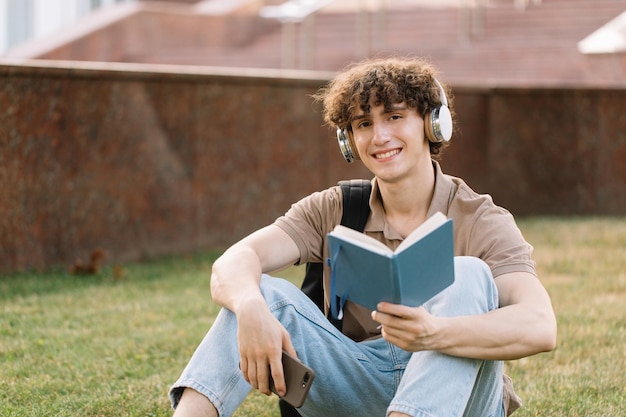 Étudiant masculin attrayant dans des écouteurs assis sur l'herbe faisant ses devoirs près de l'université le jour ensoleillé