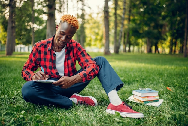 Étudiant noir dans des verres écrivant dans un cahier sur l'herbe dans le parc d'été.