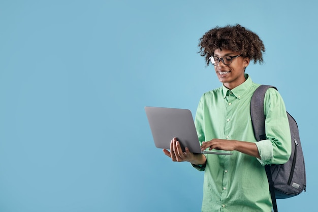 Étudiant noir positif en tenue décontractée avec sac à dos à l'aide d'un ordinateur portable moderne sur studio bleu