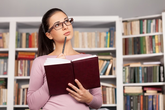 Étudiant réfléchi. Belle jeune femme tenant un livre et détournant les yeux en se tenant debout à la bibliothèque