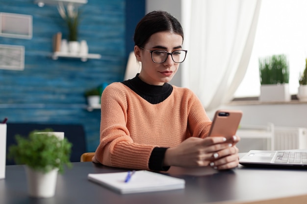 Étudiant regardant l'écran du smartphone tout en envoyant un message texte assis à la table du bureau dans le salon. Étudiant parcourant des informations sur le mode de vie sur Internet pendant la quarantaine du coronavirus