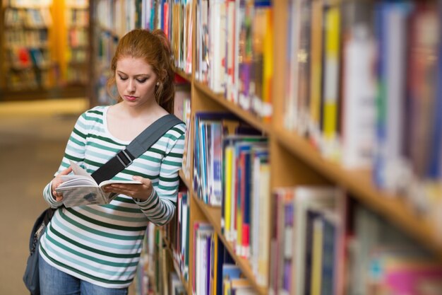 Étudiant rousse lisant livre s&#39;appuyant sur une étagère dans la bibliothèque