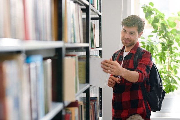 Étudiant avec sac à dos en prenant l'un des livres de l'étagère dans la bibliothèque du collège