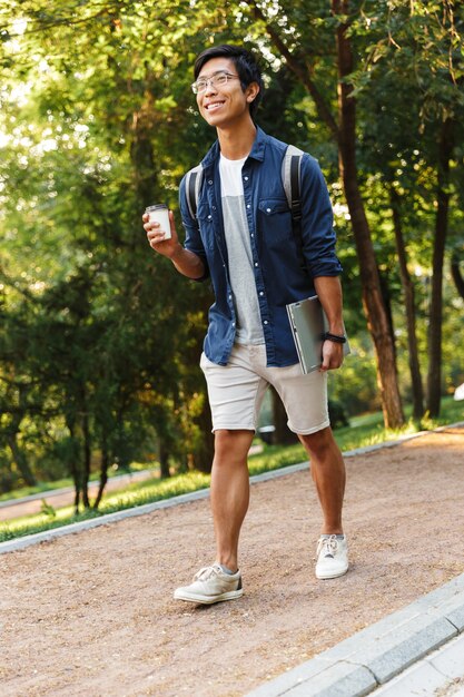 Étudiant de sexe masculin asiatique souriant vertical à lunettes marchant avec ordinateur portable dans le parc