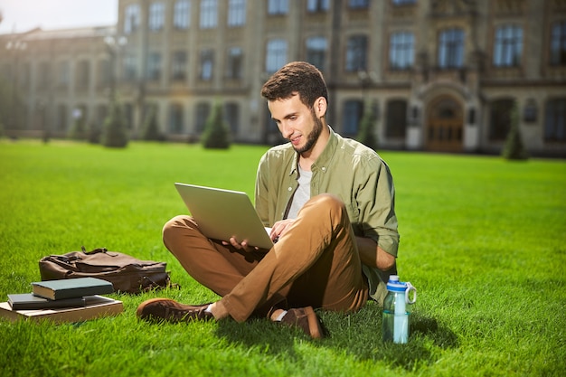 Étudiant de sexe masculin heureux souriant avec un ordinateur dans ses mains assis les jambes croisées sur l'herbe