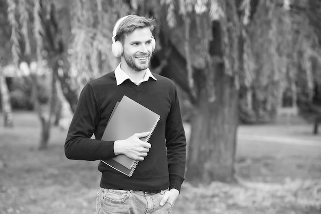 Étudiant universitaire moderne debout dans le parc écoutant de la musique avec des écouteurs et lisant la vie des étudiants