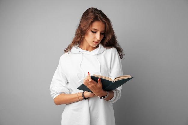 Étudiante adolescente en chandail à capuchon blanc tenant un livre dans ses mains sur un fond gris