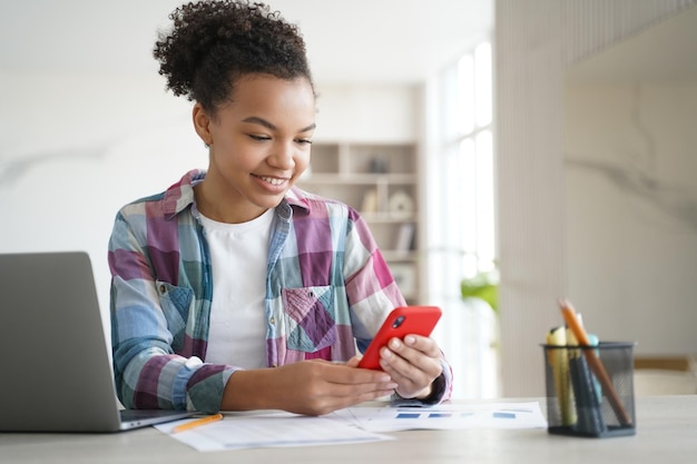 Étudiante adolescente de race mixte utilisant un smartphone discutant sur un réseau social tout en apprenant sur un ordinateur portable