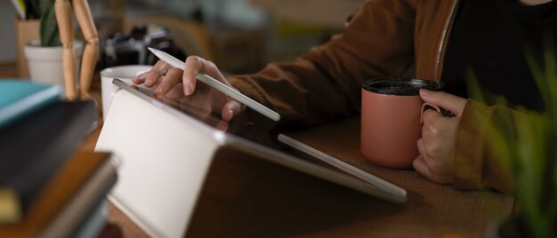 Étudiante à l'aide de tablette tout en tenant une tasse de café sur une table de travail avec pile de livres et de fournitures