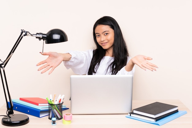 Étudiante asiatique dans un lieu de travail avec un ordinateur portable sur un mur beige présentant et invitant à venir avec la main