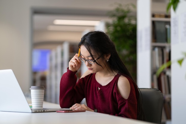 Étudiante asiatique se préparant à l'examen dans une bibliothèque publique assise à table avec un ordinateur portable