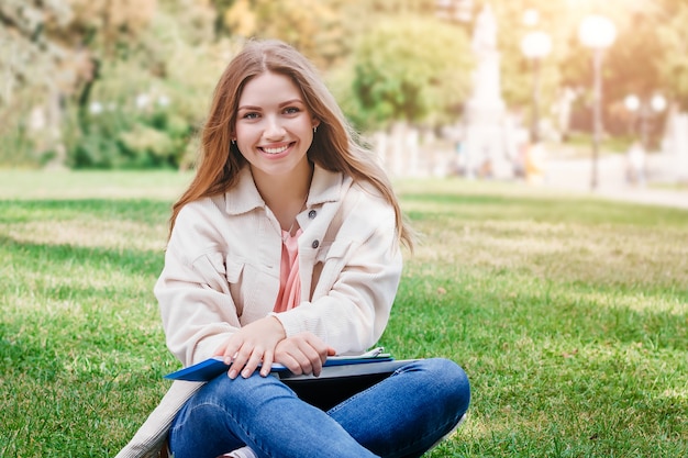 Étudiante blonde assise sur l'herbe, sourit et enseigne des leçons