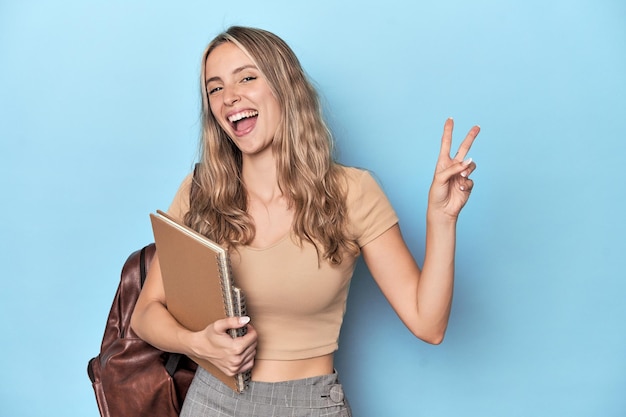 Étudiante blonde avec cahiers et sac à dos joyeuse et insouciante montrant un symbole de paix avec les doigts