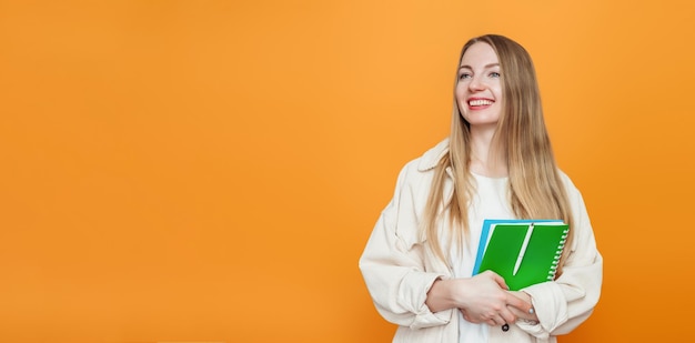 Étudiante blonde caucasienne tenant un cahier de livre souriant et regardant loin isolé sur fond orange en studio Copier l'espace