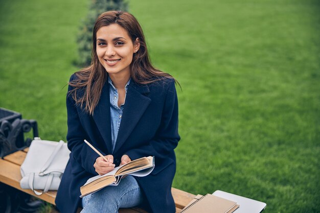 Étudiante brune élégante étudiant à l'extérieur alors qu'elle était assise sur le banc et souriant