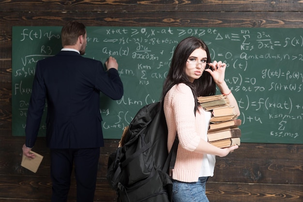 Étudiante brune avec une pile de livres debout dans la salle de classe pendant que le professeur écrit une formule sur un tableau vert
