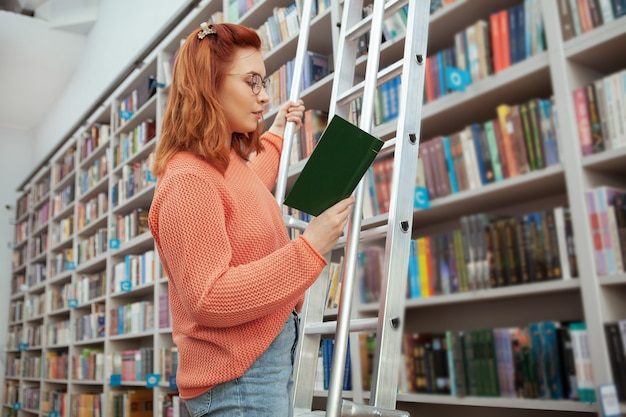 Étudiante debout sur l'échelle à la bibliothèque, lisant un livre