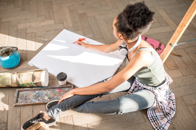 Étudiante dessinant sur la toile blanche assise sur le sol. Vue d'en-haut