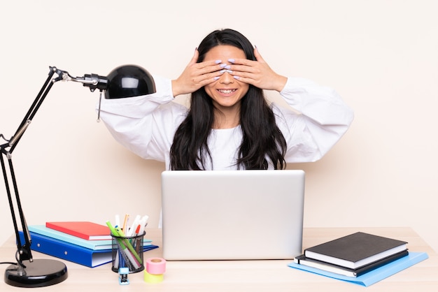 Étudiante fille asiatique dans un lieu de travail avec un ordinateur portable isolé sur un revêtement mural beige les yeux par les mains