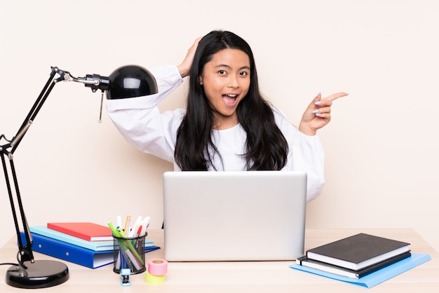 Étudiante fille asiatique dans un lieu de travail avec un ordinateur portable sur le mur beige surpris et pointant le doigt sur le côté