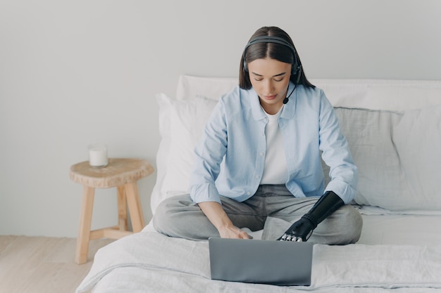 Étudiante handicapée étudiant en ligne sur un ordinateur portable à l'aide d'un bras prothétique bionique assis sur le lit