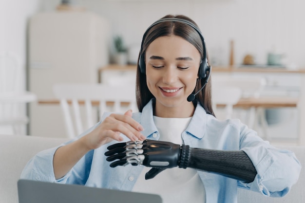 Étudiante handicapée souriante dans un casque à l'aide d'un bras prothétique bionique assis sur un ordinateur portable à la maison