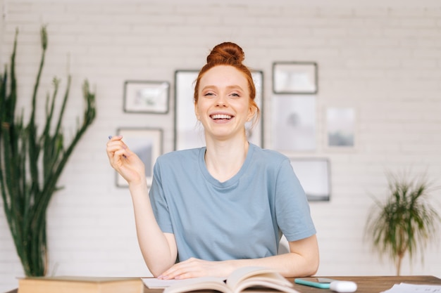 Étudiante heureuse de jeune femme rousse étonnée s'asseyant à une table avec des manuels et un classeur